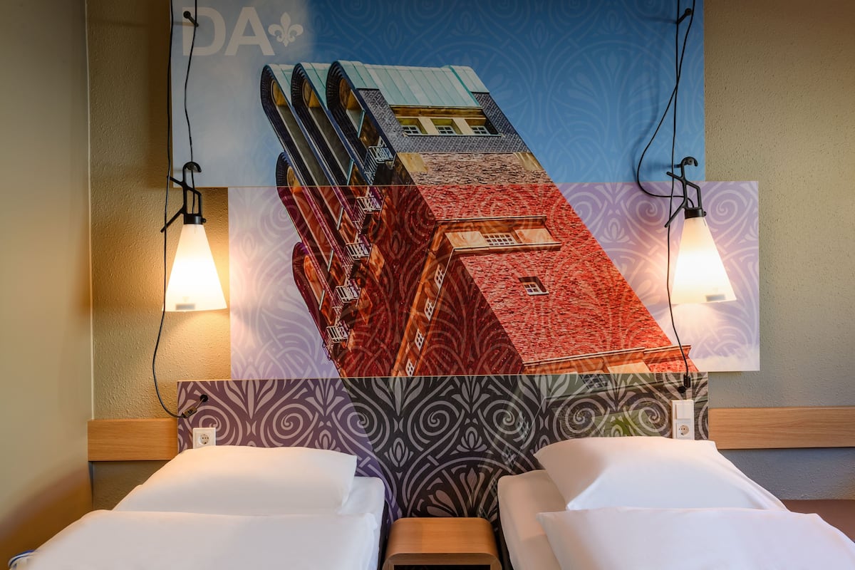 Quarto de hotel com duas camas, cabeceira estampada e uma obra de arte na parede retratando um edifício.