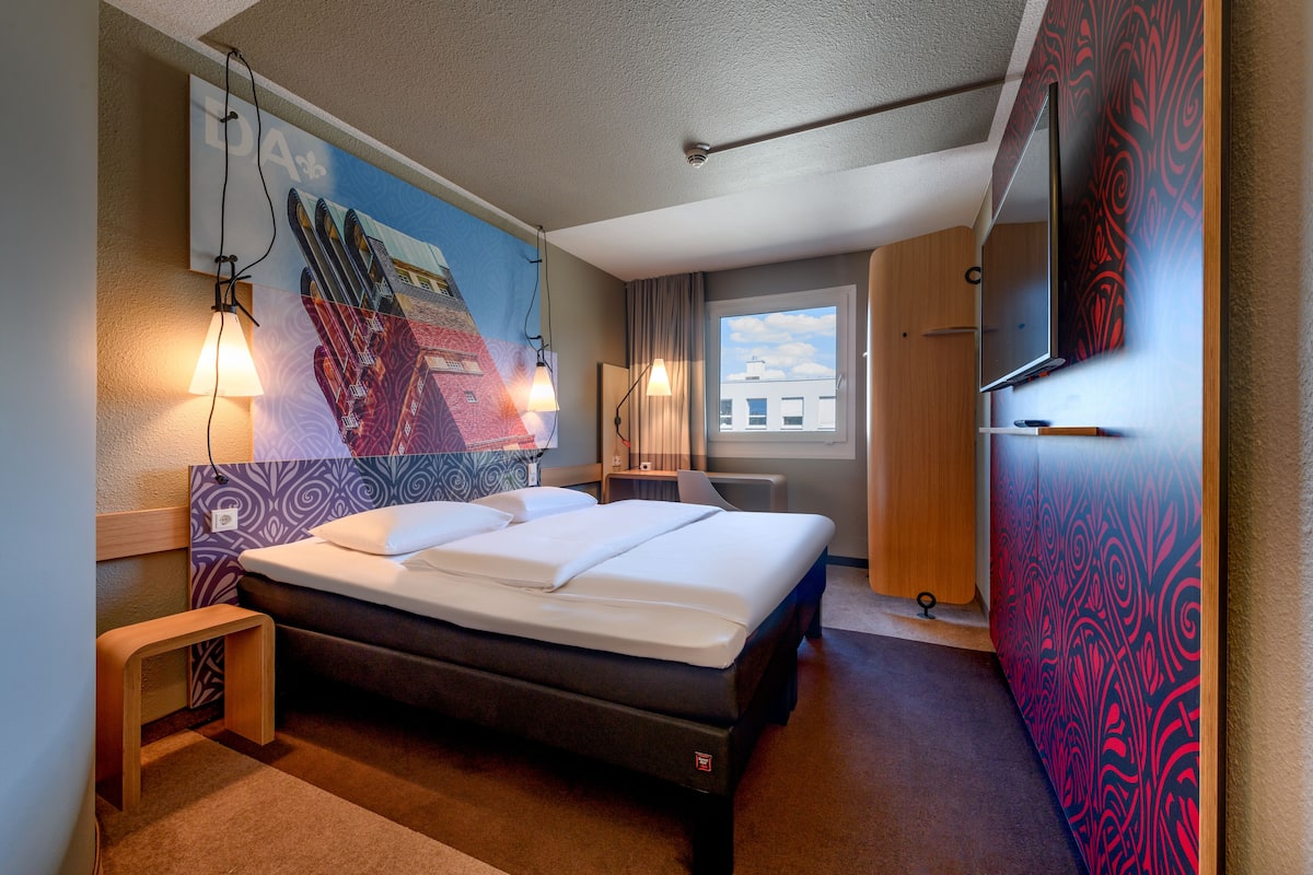 Quarto de hotel com cama, mesas de cabeceira, televisão e janela com vista para edifícios.