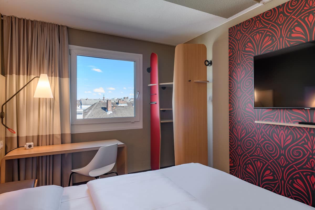 Quarto de hotel com cama, escrivaninha, cadeira, janela com vista, televisão e uma prancha de surf.