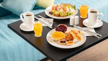 Una bandeja de desayuno con café, jugo de naranja, croissants y una fuente de frutas y verduras sobre una cama con ropa de cama azul.