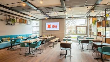 Un ufficio moderno con un ampio schermo che mostra il logo "ibis", tavoli e sedie in legno e una vista all'esterno attraverso ampie finestre.