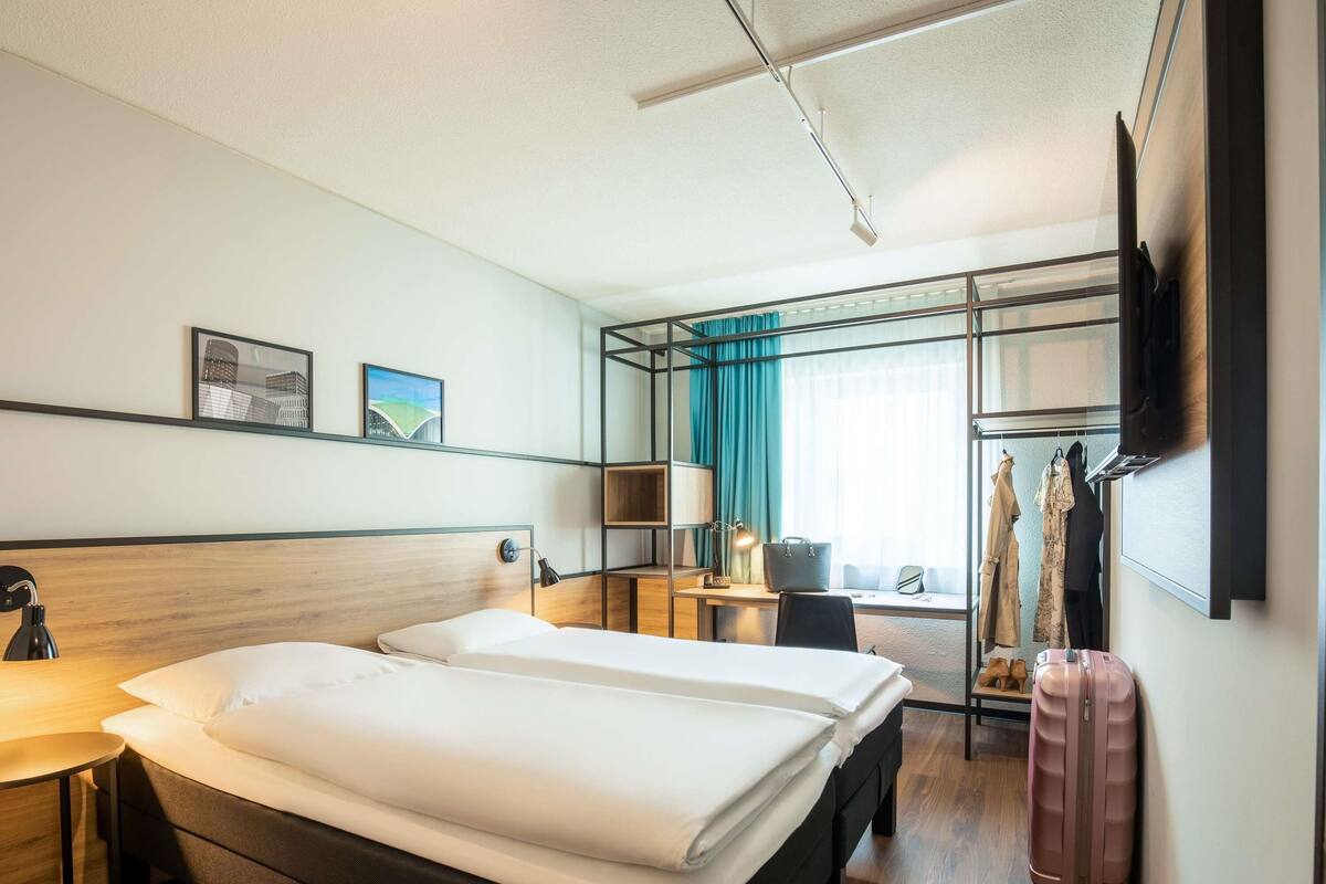 A hotel room with two beds, a desk, a chair, a TV, and a suitcase.