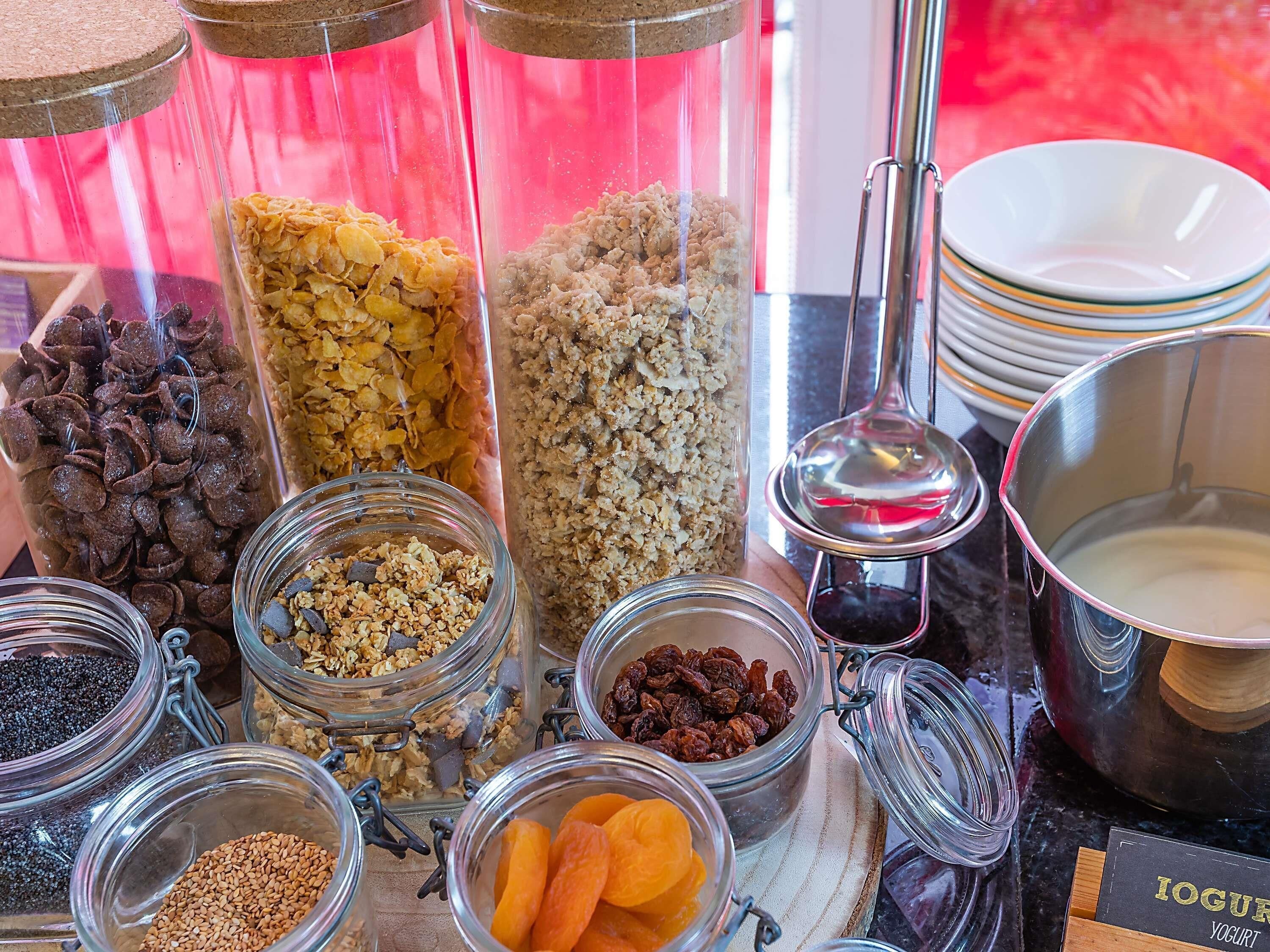A variety of food items in glass containers, including granola, dried fruits, and nuts, with a bowl of yogurt and a spoon nearby.