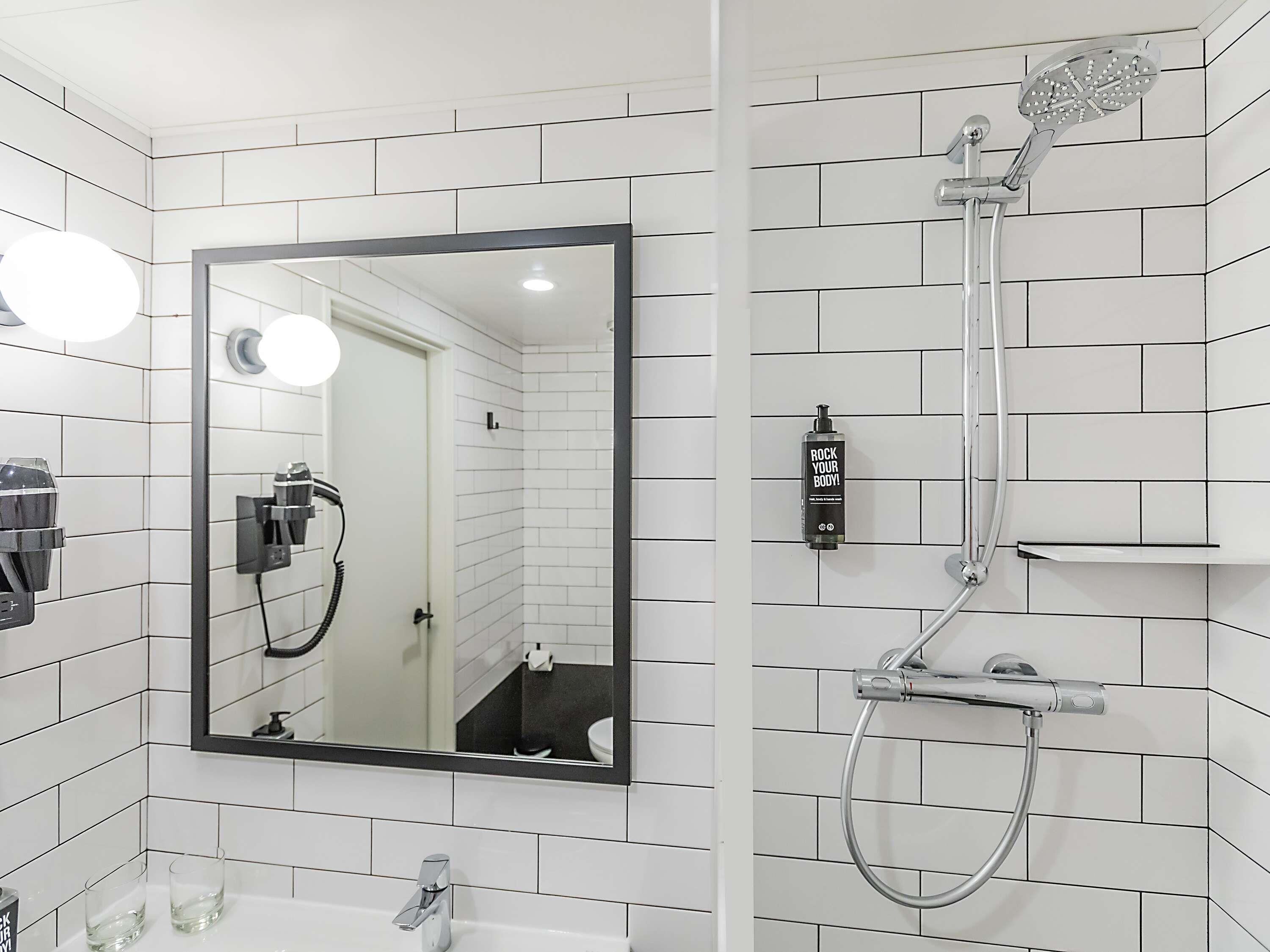 Un baño con paredes de azulejos blancos, un espejo grande, ducha con cabezal de ducha de mano y una botella de champú.