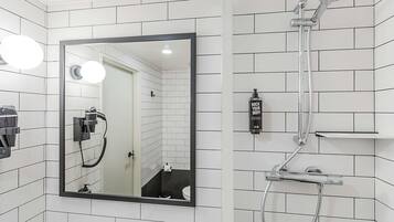 A bathroom with white tiled walls, a large mirror, a shower with a handheld showerhead, and a bottle of shampoo.