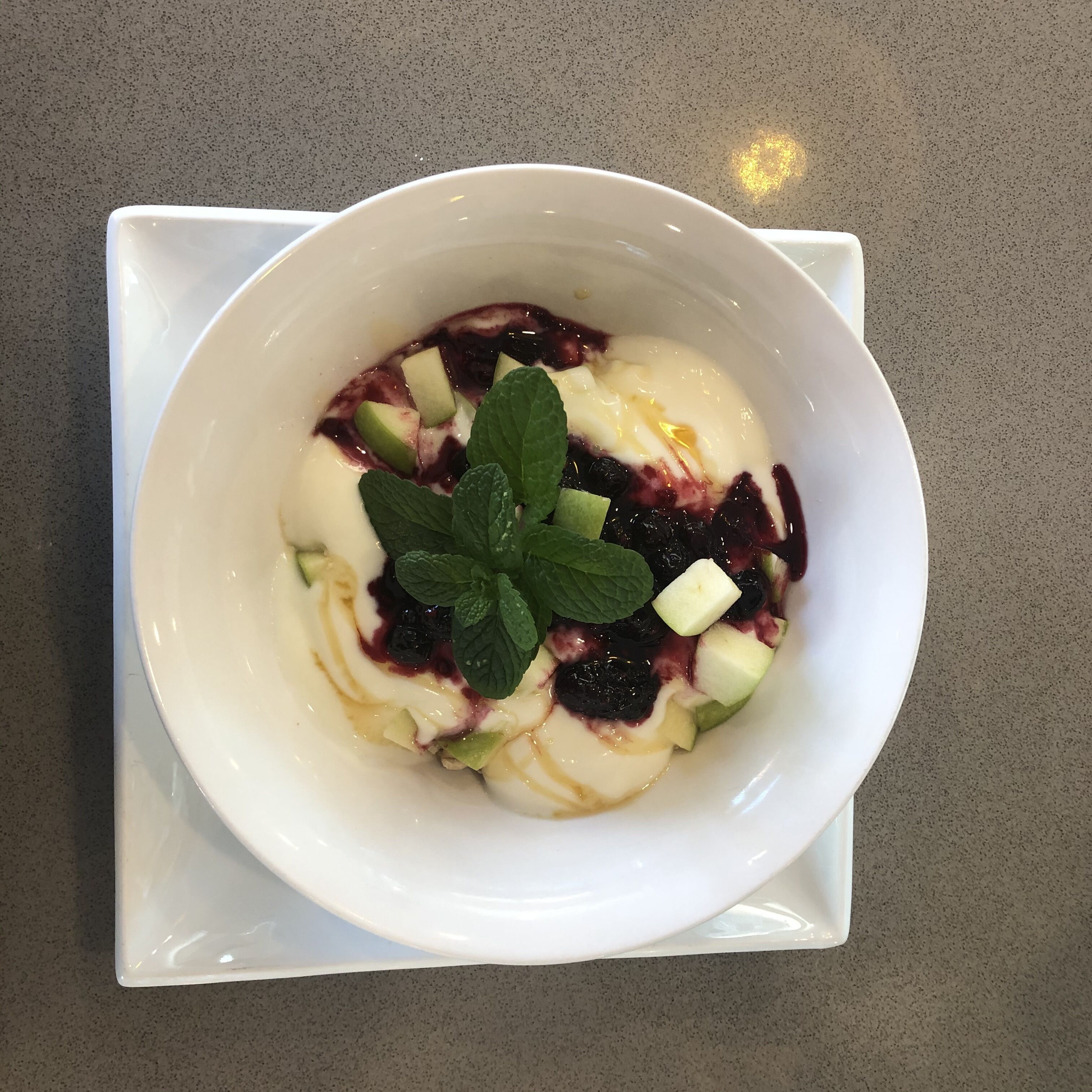 En vit skål med efterrätt, garnerad med frukt och mynta, står på en bänkskiva med fläckigt mönster.