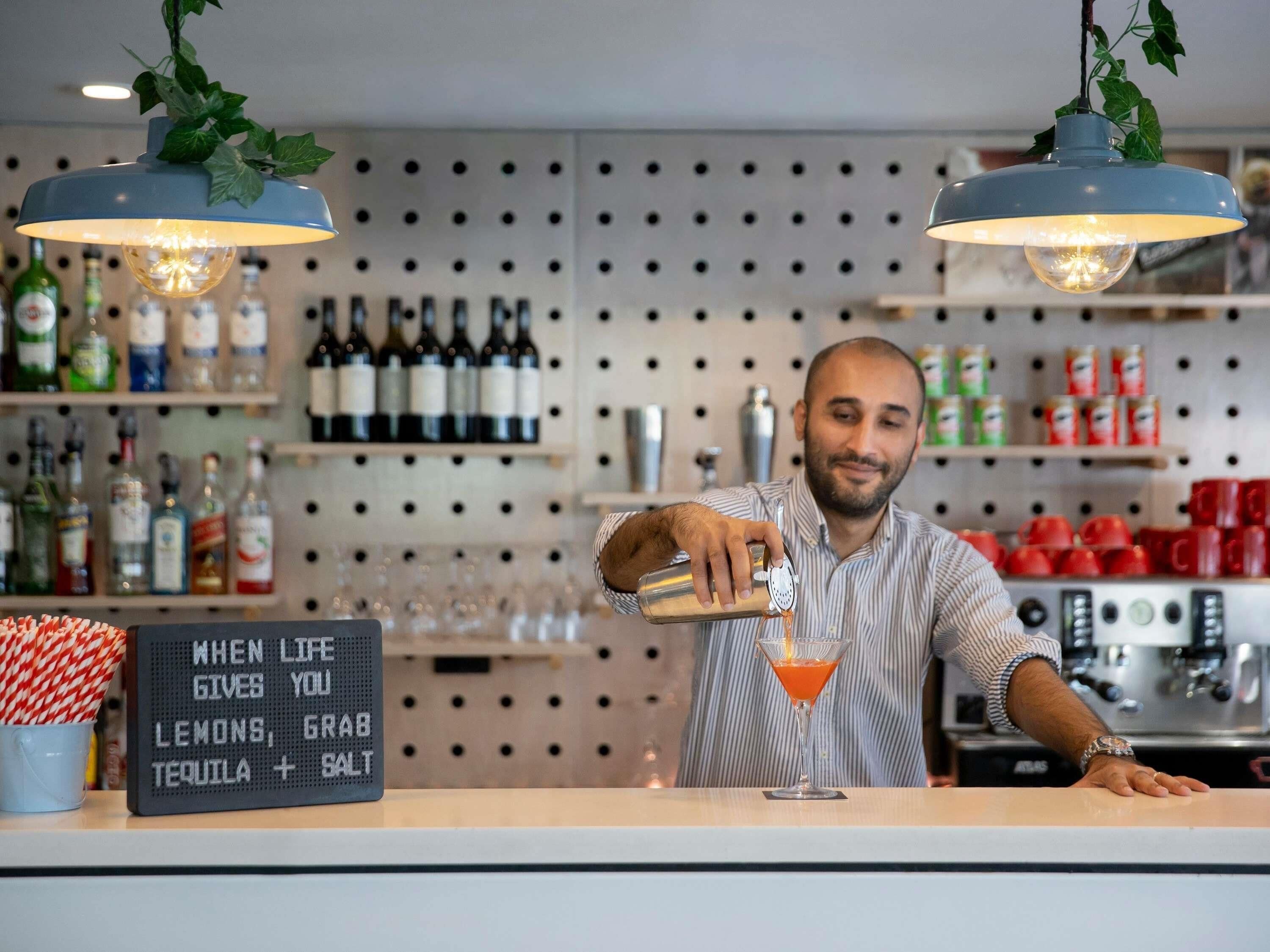 A bar area with a bartender pouring a drink into a martini glass, shelves with bottles, and a sign with a humorous message.