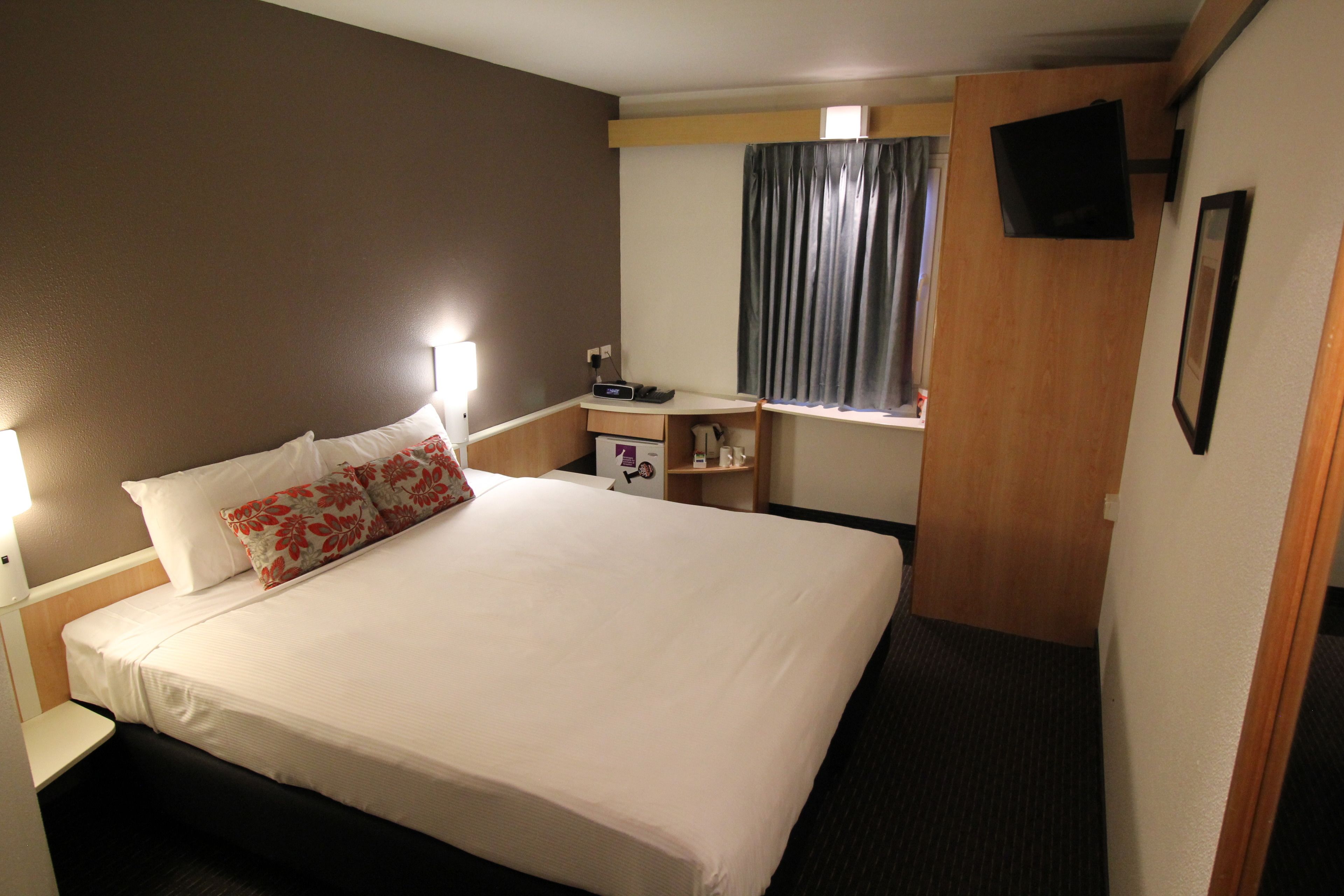 Een hotelkamer met een bed, nachtkastje, lamp, televisie en een raam met gordijnen.