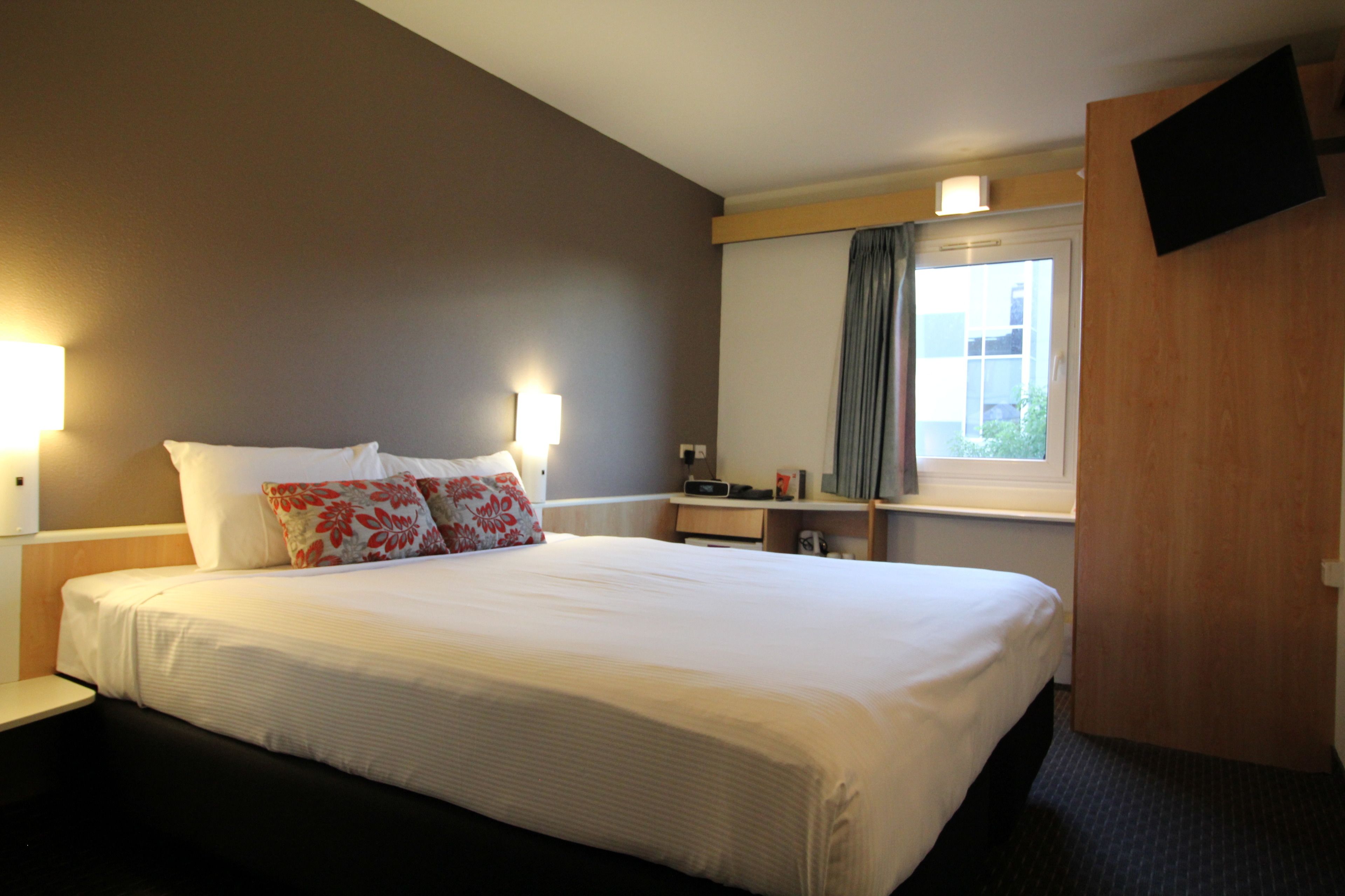 A hotel room with a large bed, bedside tables, a flat-screen TV mounted on the wall, and a window with curtains.