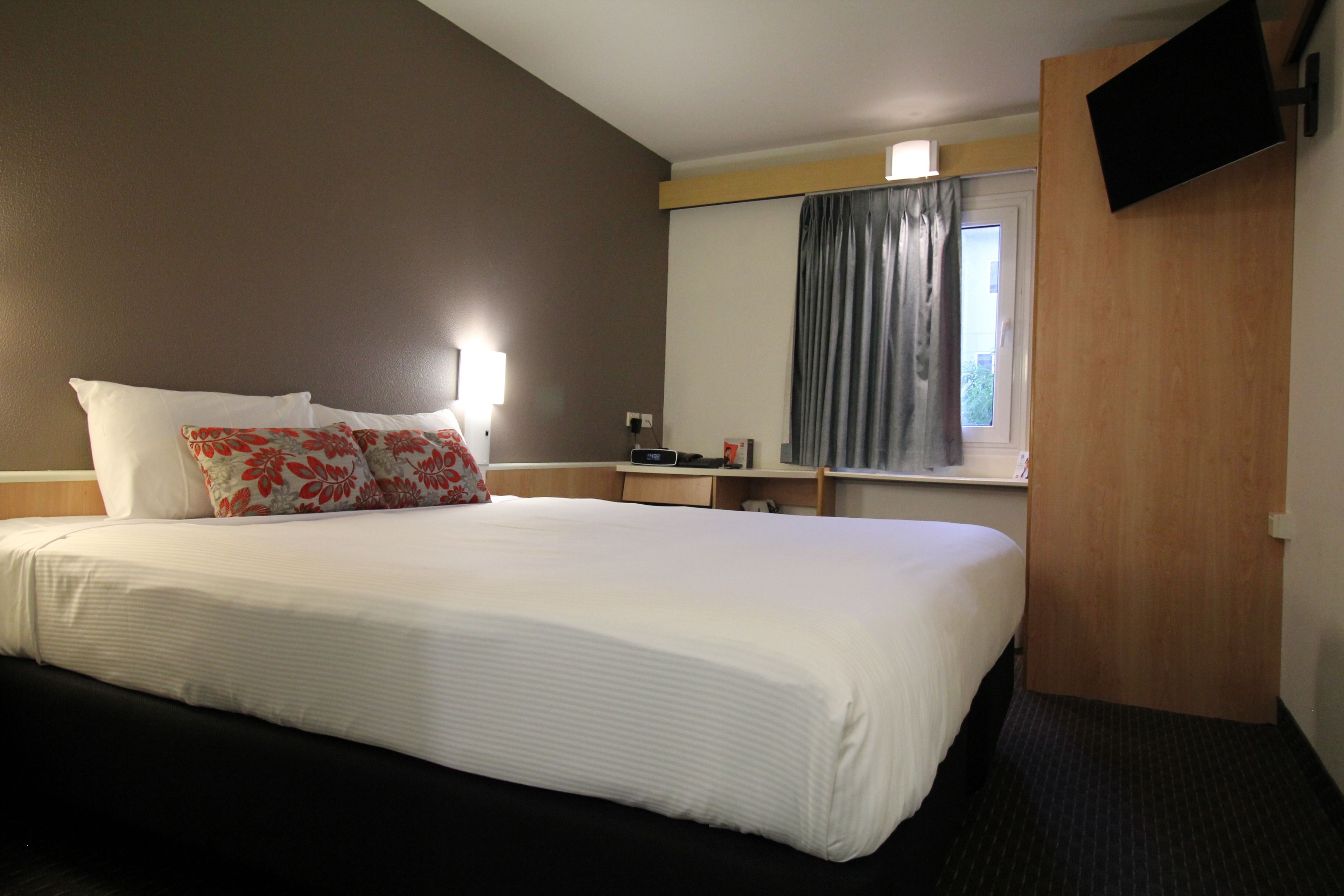 Een hotelkamer met een groot bed, een flatscreen televisie aan de wand, een nachtkastje met een lamp en een raam met gordijnen.