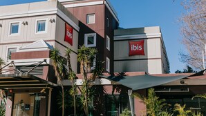 Un hotel de varios pisos con letrero 'ibis', un acceso cubierto y palmeras.