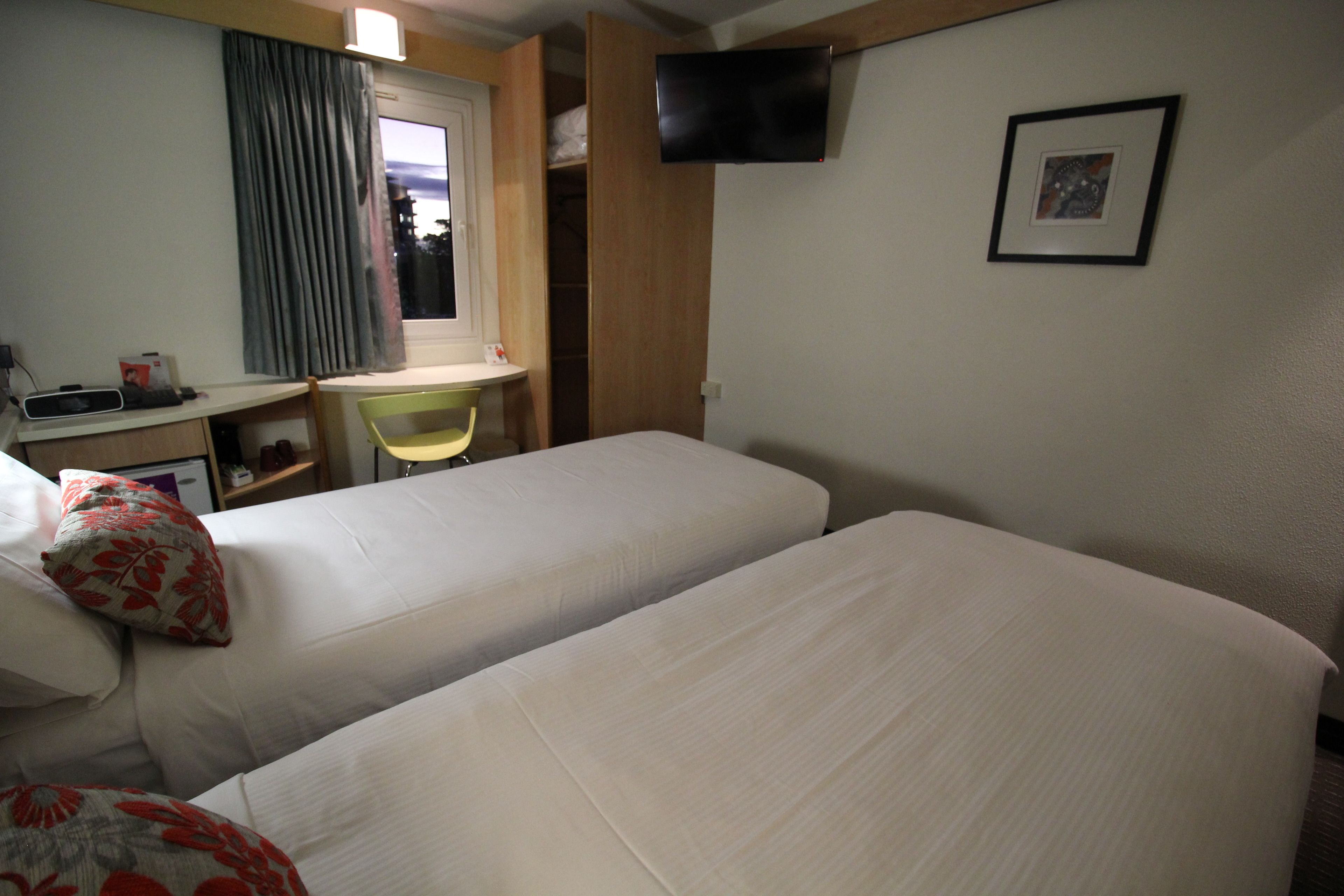 A hotel room with two beds, a desk, a chair, a TV mounted on the wall, and a window with curtains.