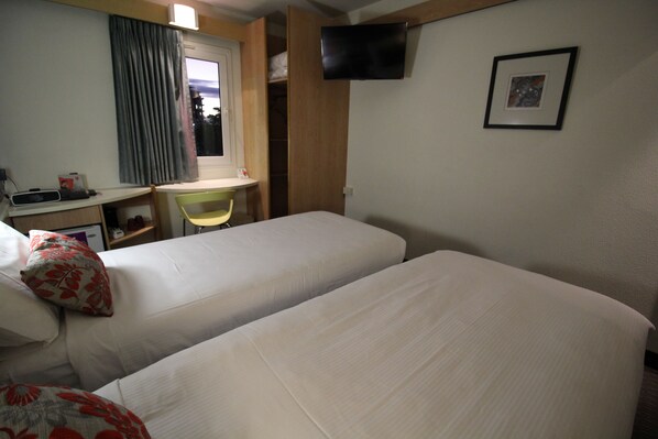 Quarto de hotel com duas camas, uma escrivaninha, uma cadeira, televisão fixada na parede e uma janela com cortinas.