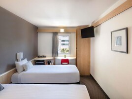 A hotel room with two beds, a desk with a chair, a TV mounted on the wall, and a window with curtains.