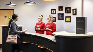 A reception desk with two staff members in red shirts, a guest with a handbag, and framed artwork on the wall. - ibis Northampton Centre (Northampton)