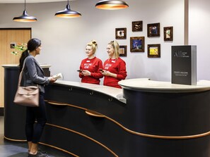 A reception desk with two staff members in red shirts, a guest with a handbag, and framed artwork on the wall. - ibis Northampton Centre (Northampton)