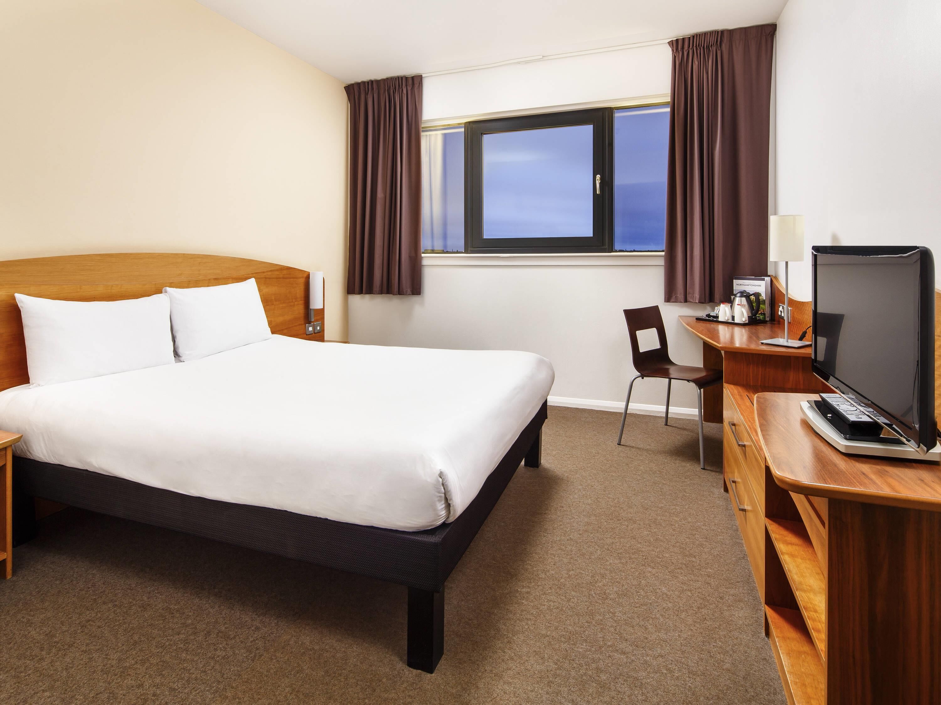 Une chambre d’hôtel avec un lit, un bureau avec un ordinateur, une chaise et une fenêtre avec des rideaux.