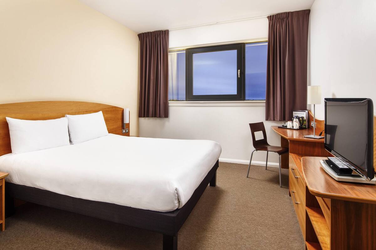Une chambre d’hôtel avec un lit, un bureau avec un ordinateur, une chaise et une fenêtre avec des rideaux.