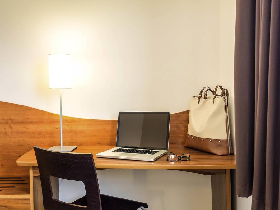 A hotel room with a wooden desk, a laptop, a pair of glasses, and a handbag.