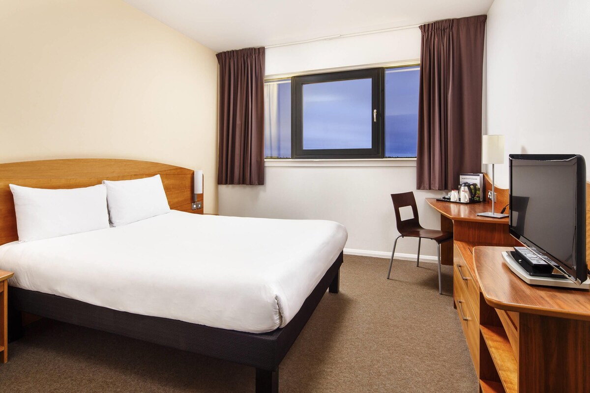 Una habitación de hotel con cama, escritorio, computadora, silla y ventana con cortinas.