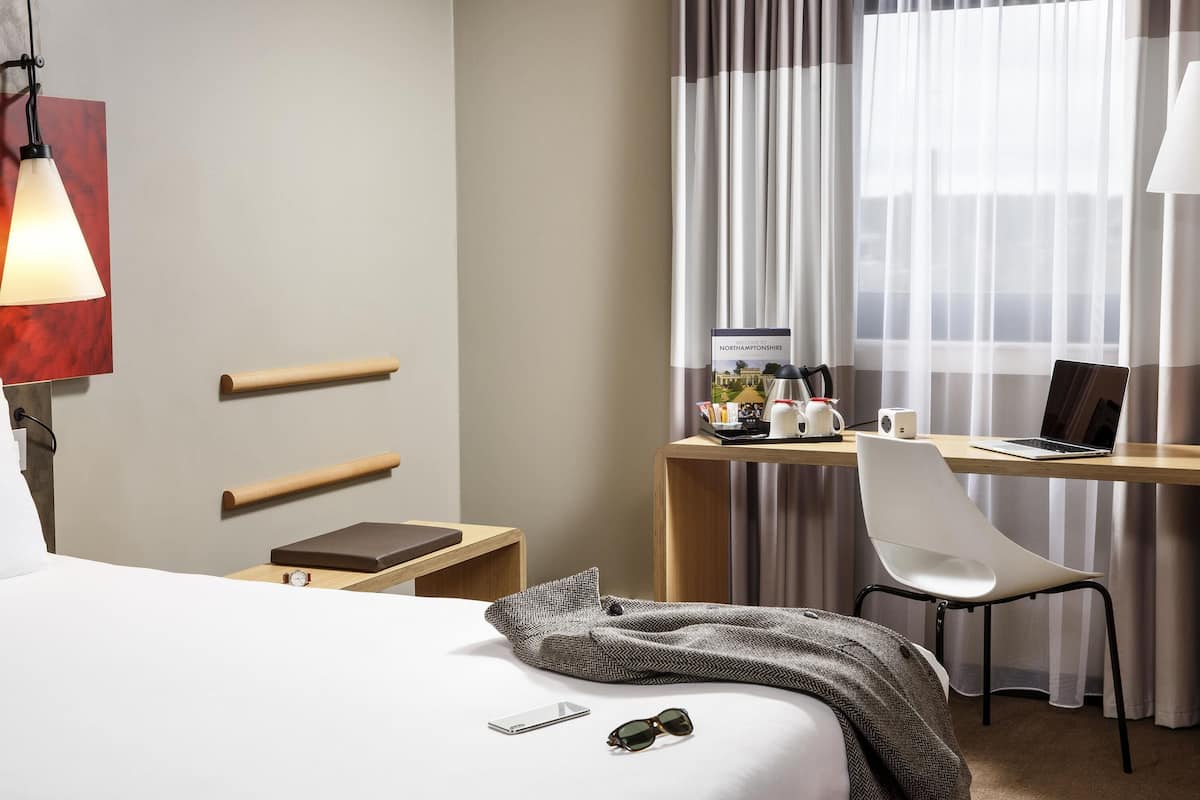 Ein Hotelzimmer mit einem Bett, einem Schreibtisch mit einem Laptop, einem Sessel, einem Nachttisch mit einer Lampe und einem Willkommensschild.