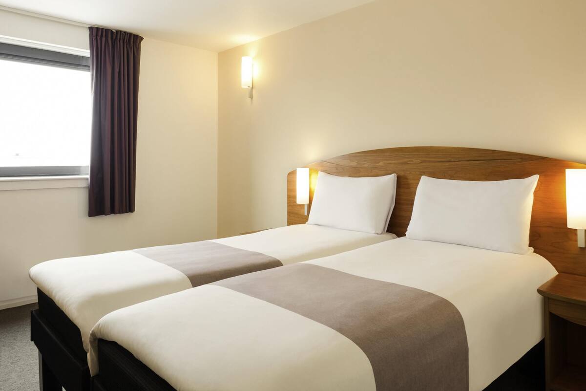 Ein Hotelzimmer mit zwei Betten, einem fensterbepflanzten Fenster und Wandleuchten.