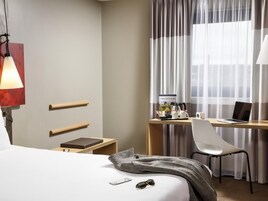 Habitación de hotel con cama, mesita de noche, lámpara, escritorio con una computadora portátil, silla y cortinas.