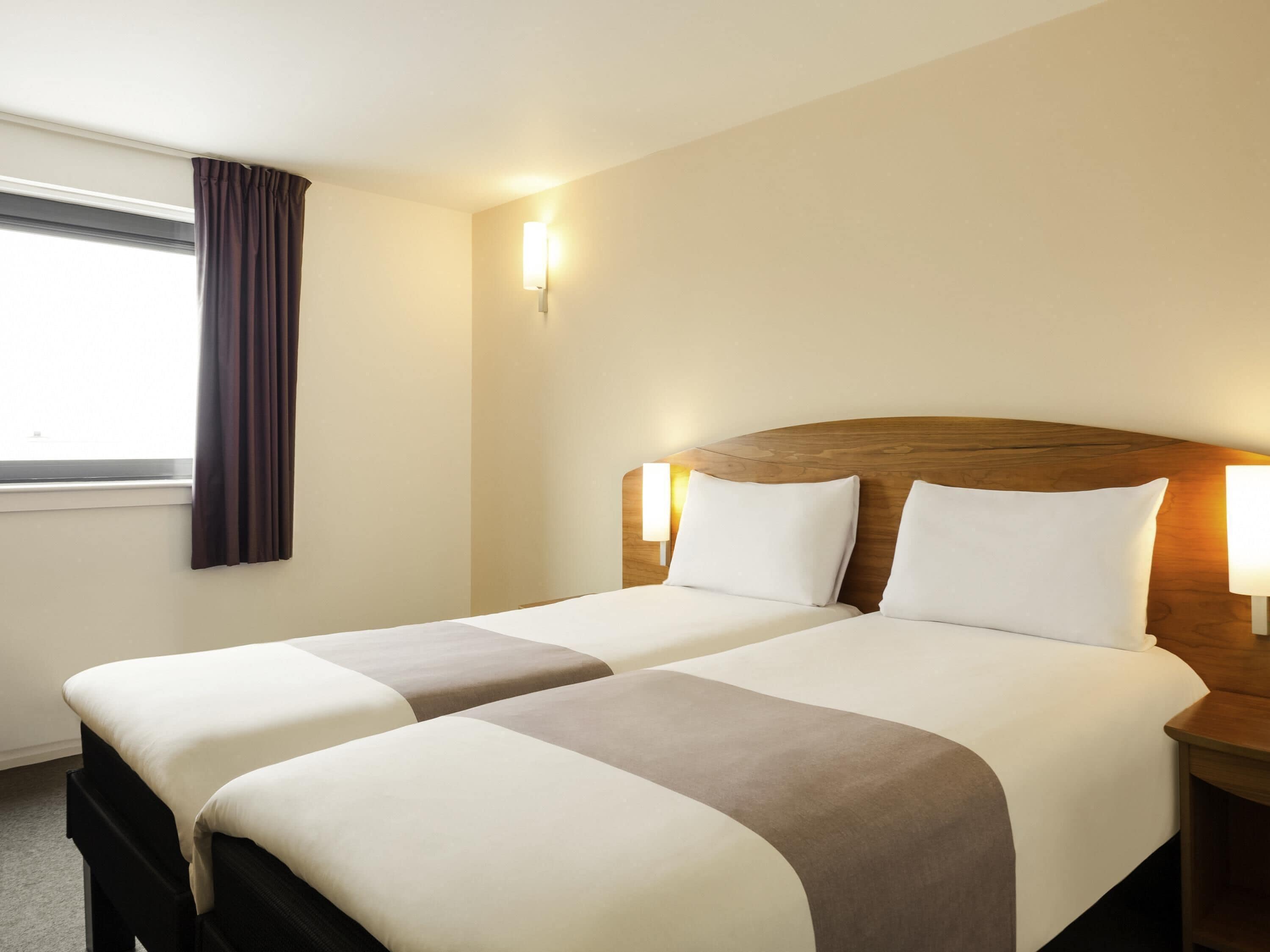 A hotel room with two beds, a window with curtains, and wall-mounted lamps.