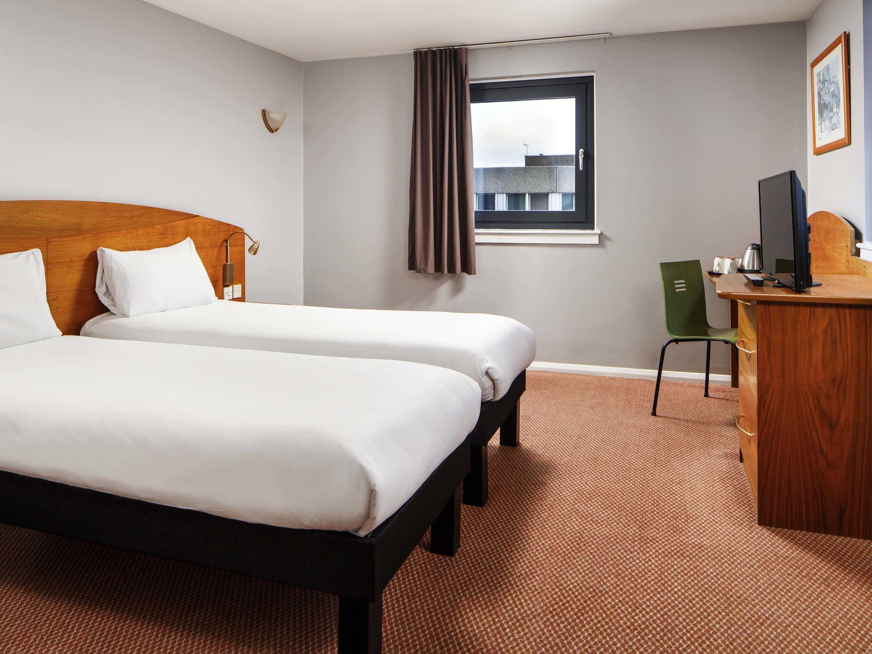 Une chambre d’hôtel avec deux lits, un bureau, une chaise, une fenêtre avec des rideaux et une lampe fixée au mur.