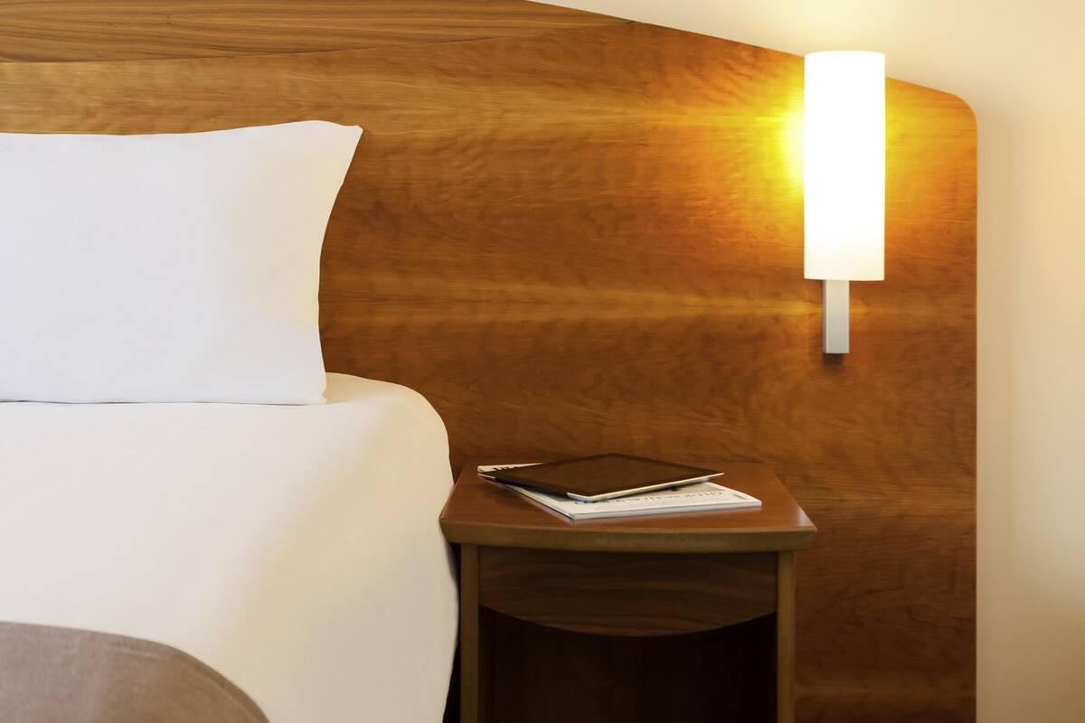 Ein ordentlich gemachtes Bett mit einem weißen Kopfkissen und einem Nachttisch mit einem Tablet und einem Buch.