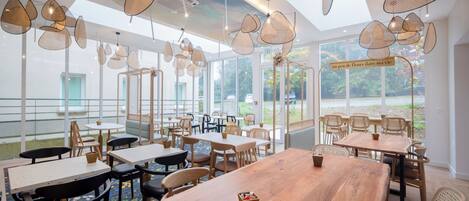 A spacious indoor dining area with multiple tables and chairs, large windows offering views of the outdoors, and hanging pendant lights with a woven design.