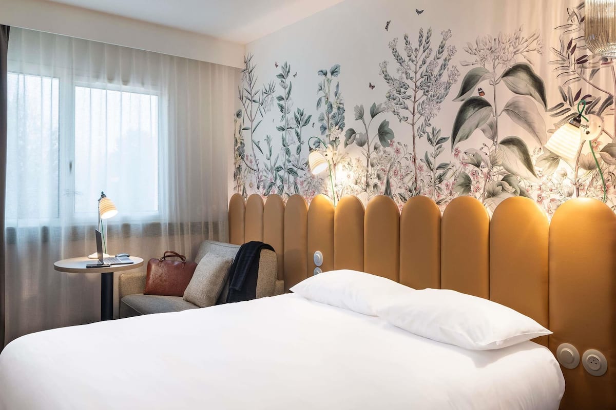 A hotel room with a large bed, a small round table with a lamp, a chair, and a decorative wall mural.
