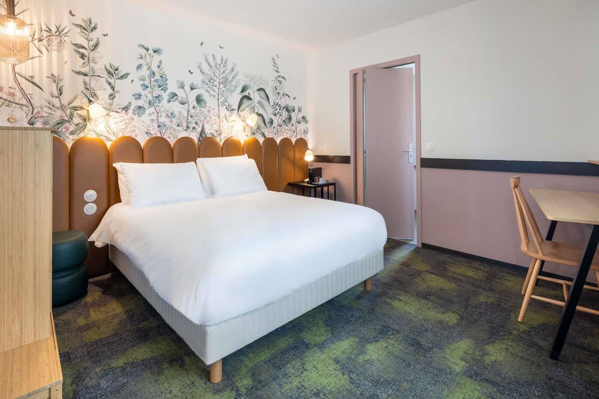 A hotel room with a bed, bedside table, lamp, and a wall with a floral mural.