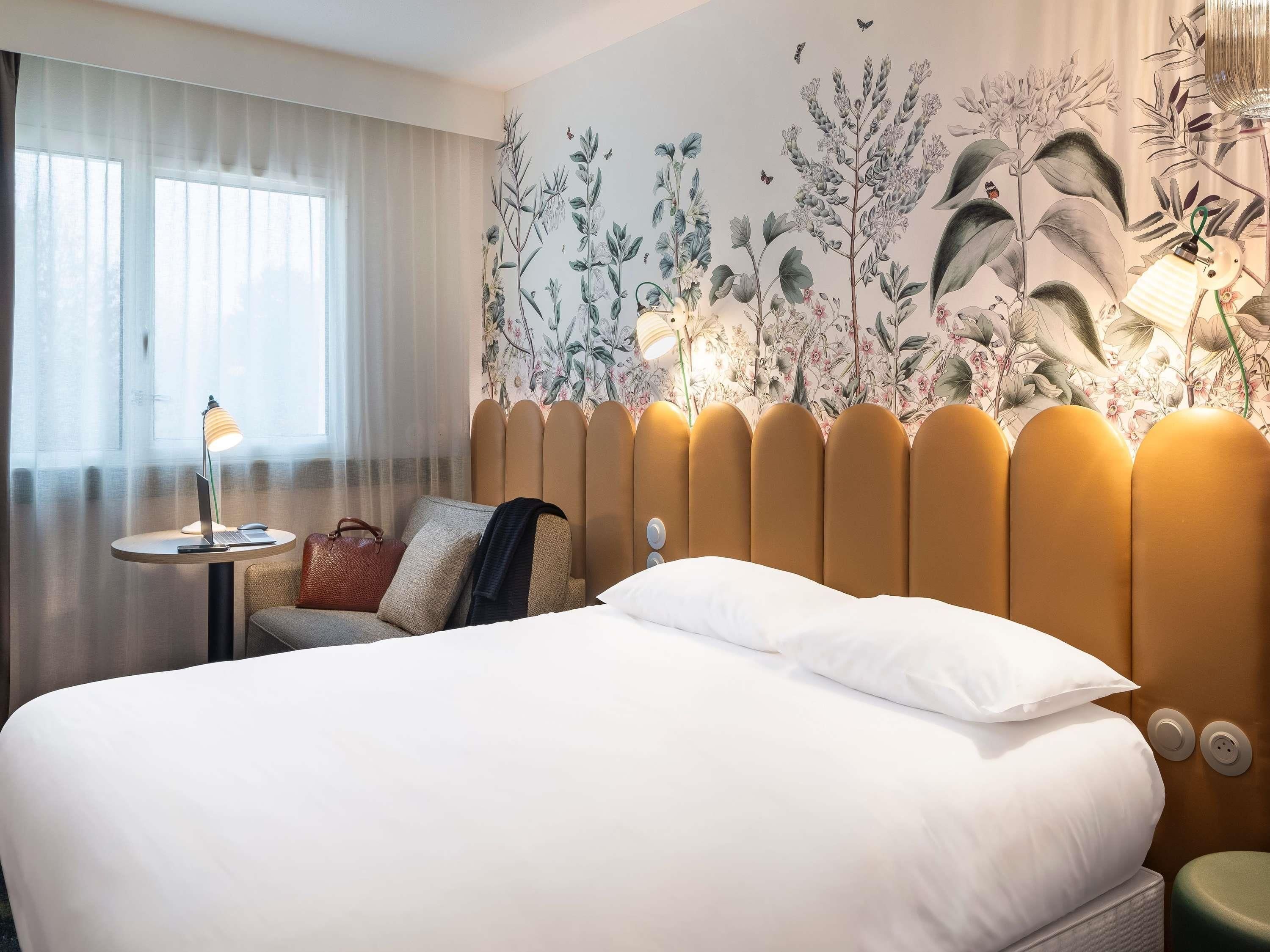 A hotel room with a bed, a round table with a lamp, a chair, and a wall with a floral mural.