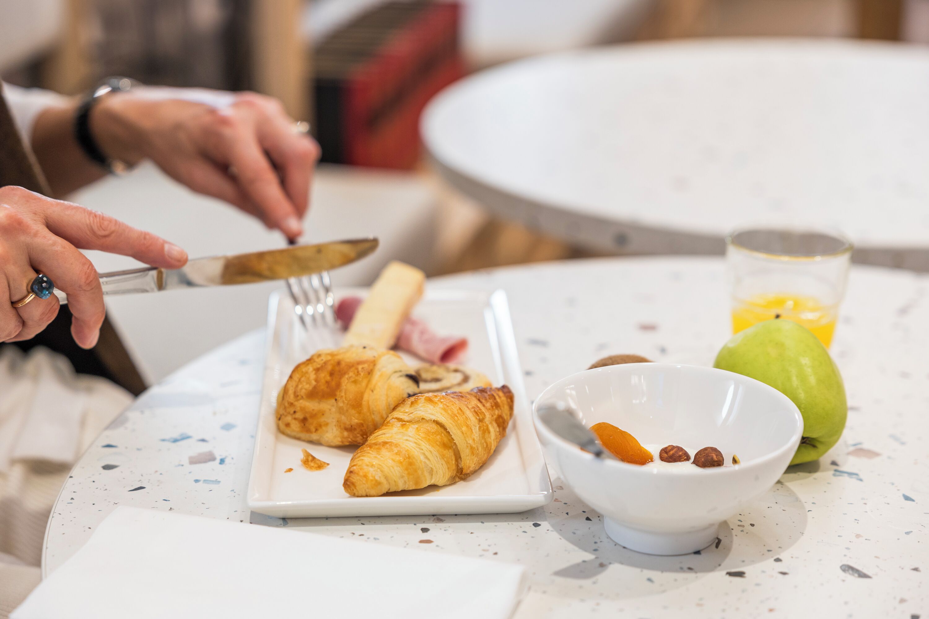 Un desayuno con croissants, un bol de nueces y fruta seca, y un vaso de zumo de naranja sobre una mesa con motas.