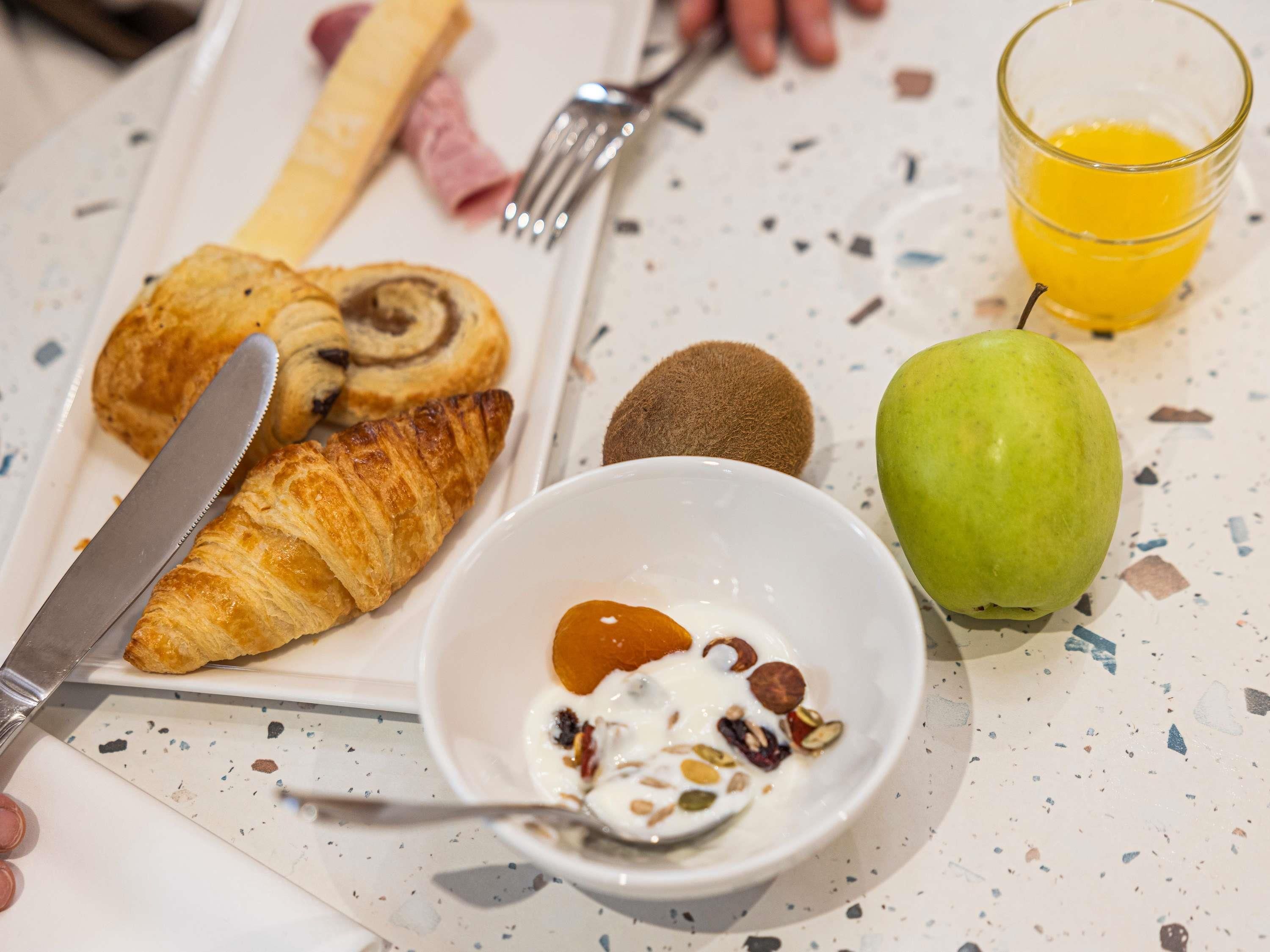 A breakfast setting with croissants, a kiwi, an apple, a glass of orange juice, and a bowl of yogurt with nuts.