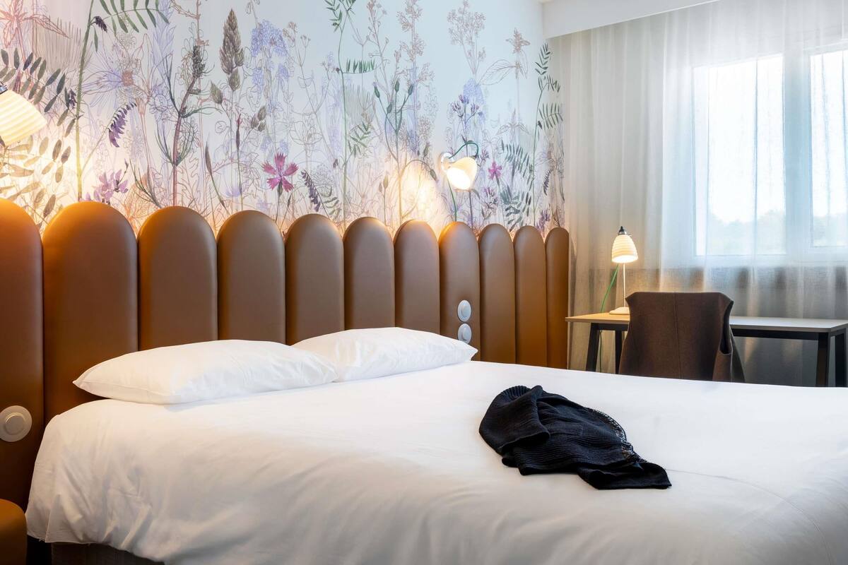 A hotel room with a bed, a desk, and a chair. There is a floral mural on the wall and a window with curtains.