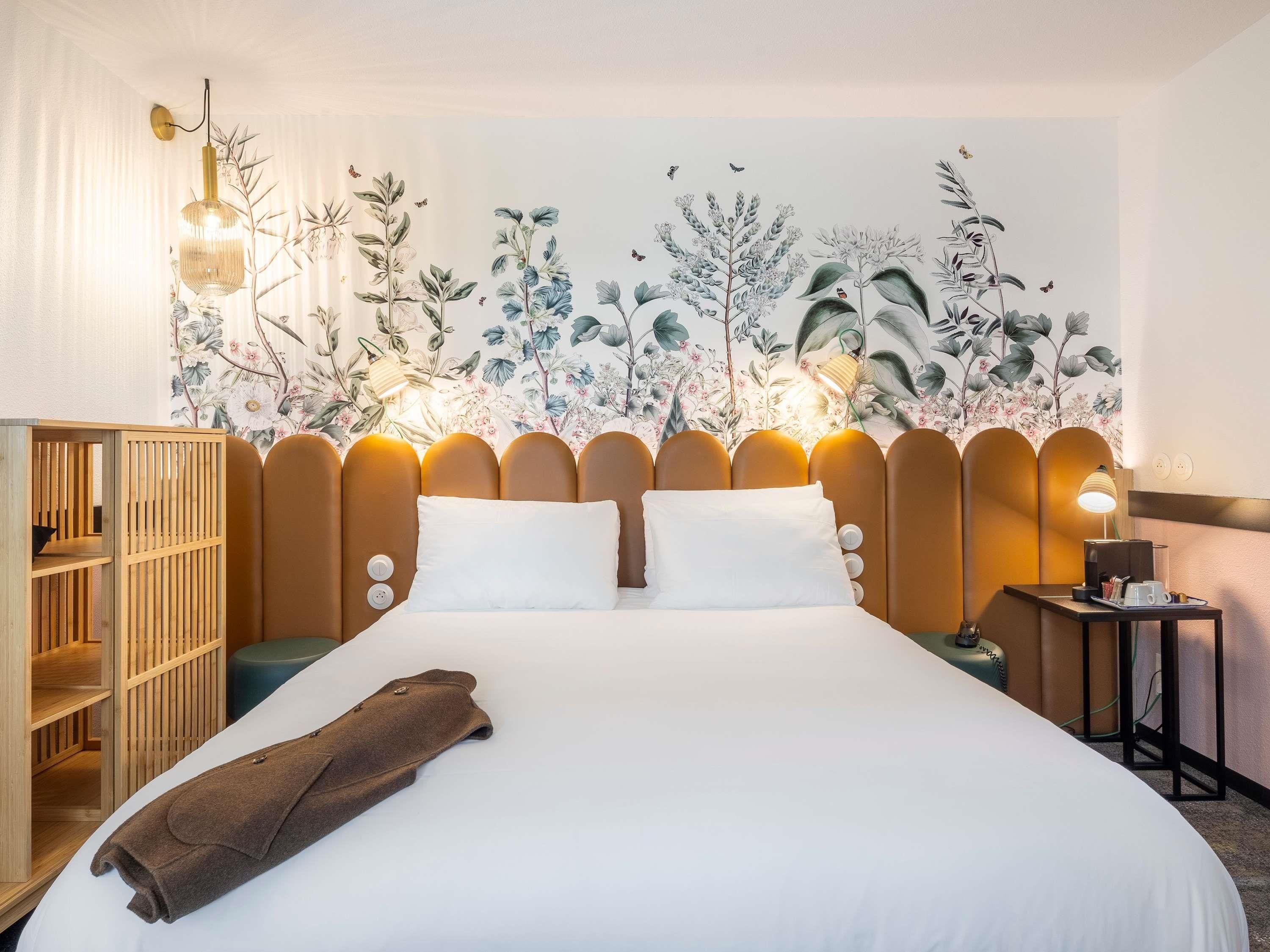 A hotel room with a large bed, a wooden wardrobe, a nightstand with a lamp, and a floral wall mural.