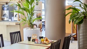 A dining area with a table set for breakfast, including croissants, a muffin, and a drink. There are chairs around the table, and a large potted plant is visible. - B&B HOTEL Paris Nord 2 CDG Aéroport (Roissy-en-France)