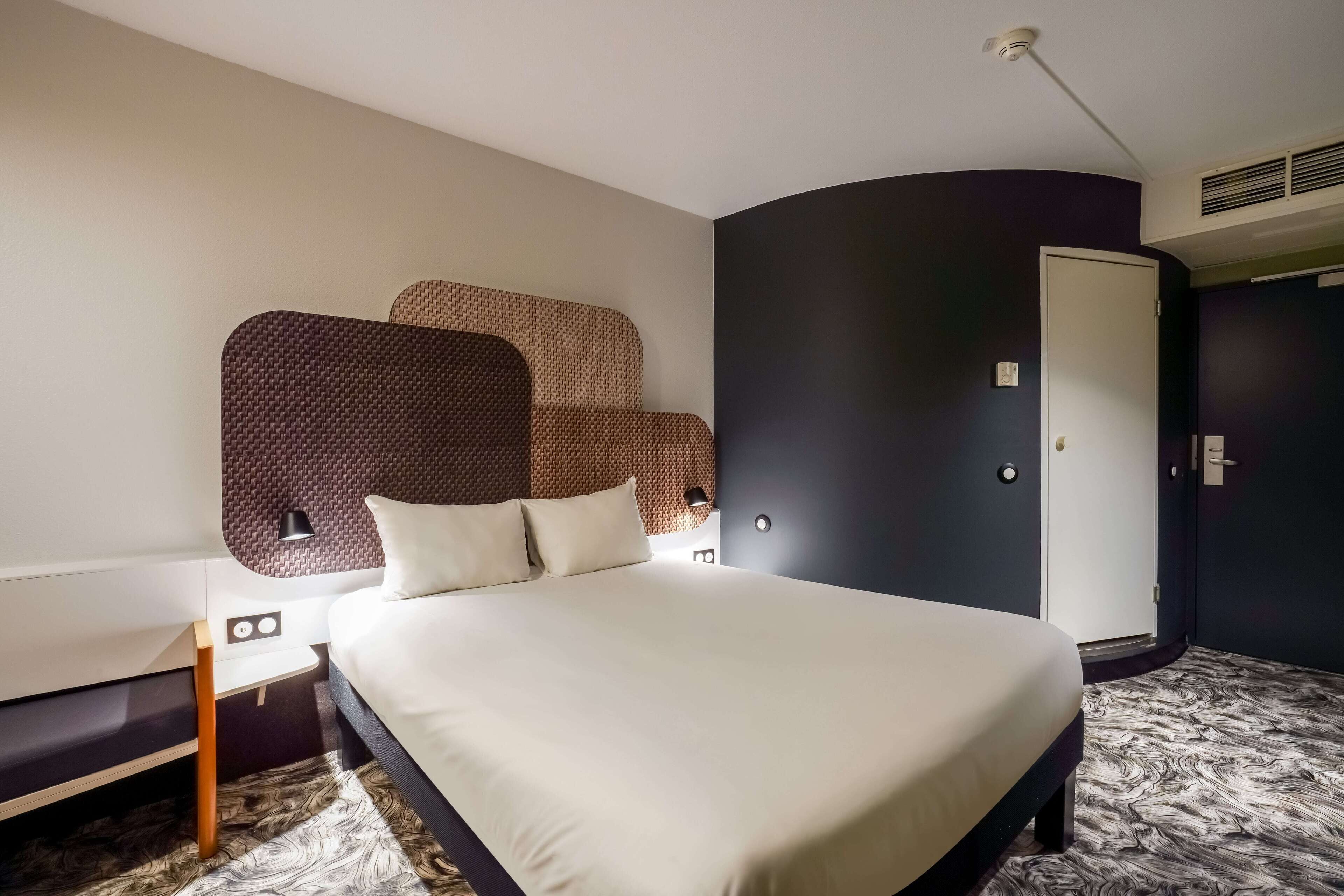 Una habitación de hotel moderna con una cama grande, una mesita de noche, una lámpara de pared y un armario.