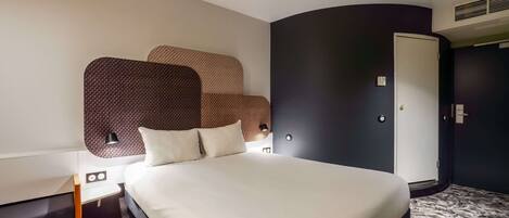 Una habitación de hotel moderna con una cama grande, una mesita de noche, una lámpara de pared y un armario.