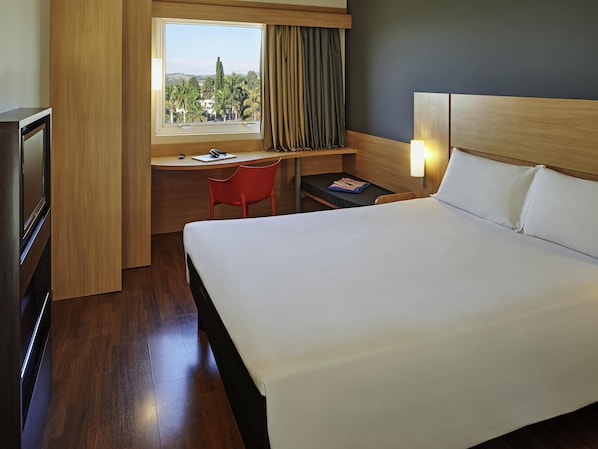 A hotel room with a bed, a desk, a chair, a TV, and a window with a view of trees. - ibis Sorocaba (Sorocaba)