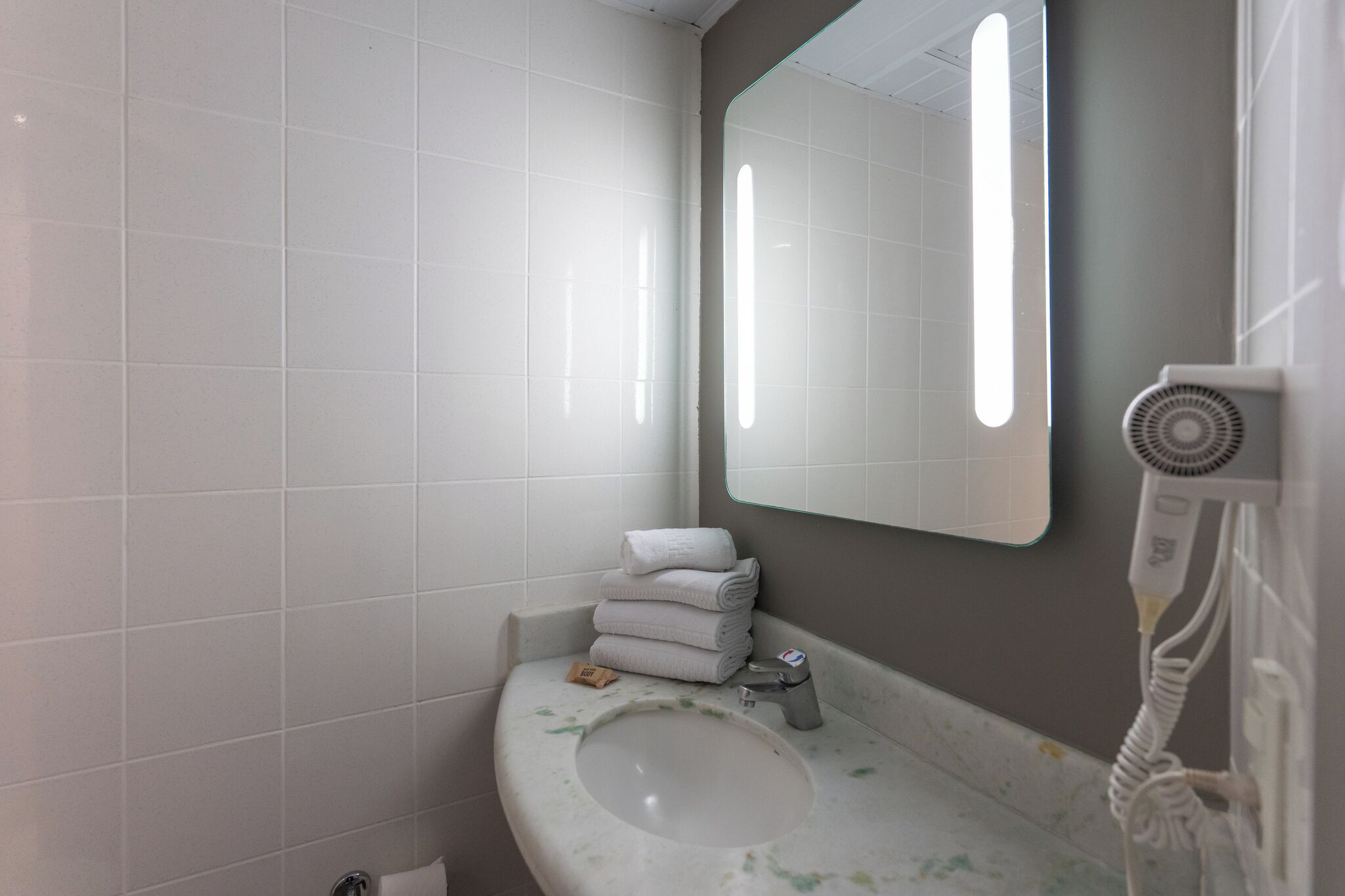 A bathroom with white tiles, a sink, a mirror, and a hairdryer.