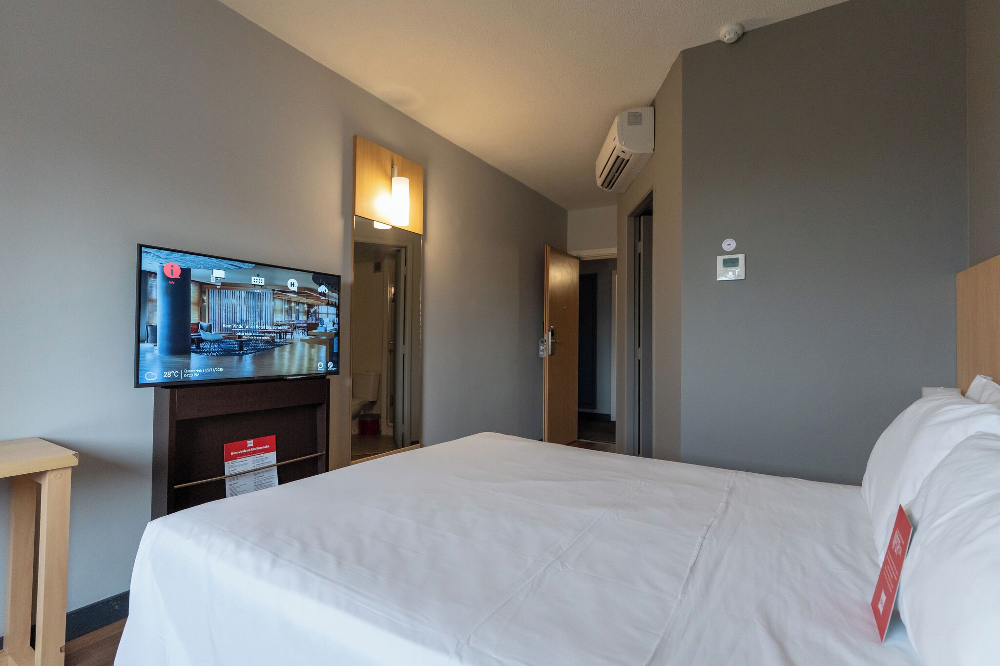A hotel room with a bed, a television mounted on the wall, a wooden nightstand, and a door leading to another room.