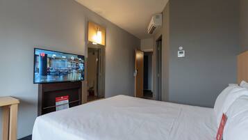 A hotel room with a bed, a television mounted on the wall, a wooden nightstand, and a door leading to another room.