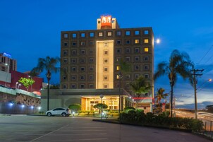 Un hotel de varios pisos con un letrero destacado de 'ibis', un estacionamiento y palmeras.