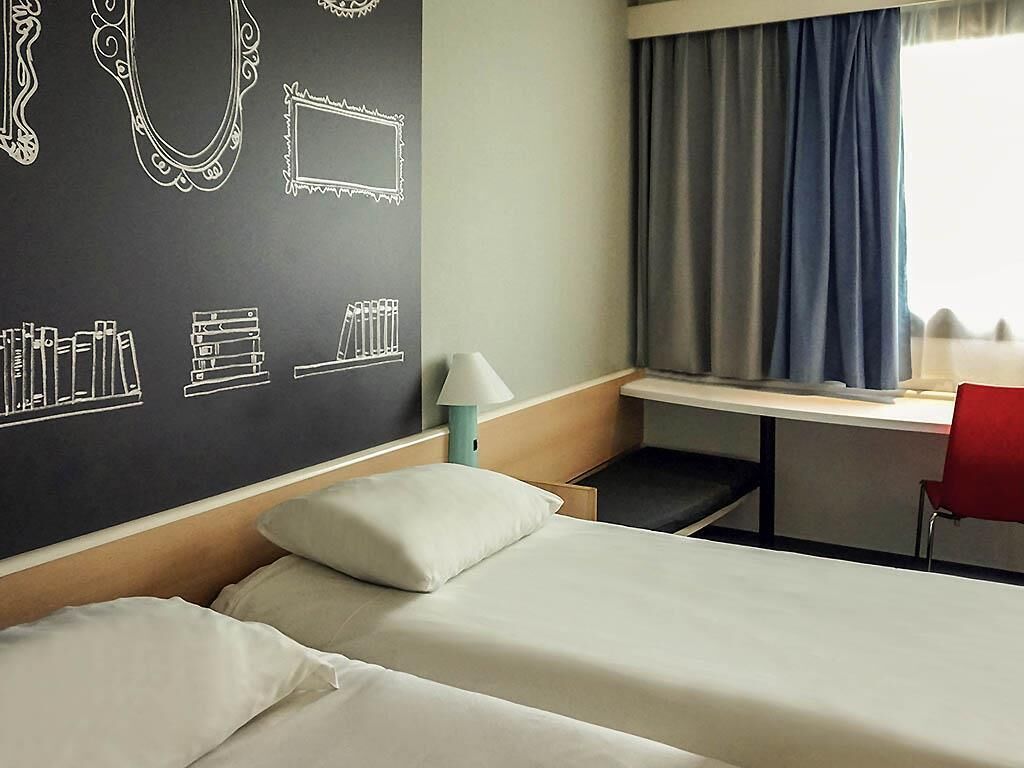 Een hotelkamer met een bed, een bureau met stoel, een lamp en een raam met gordijnen.