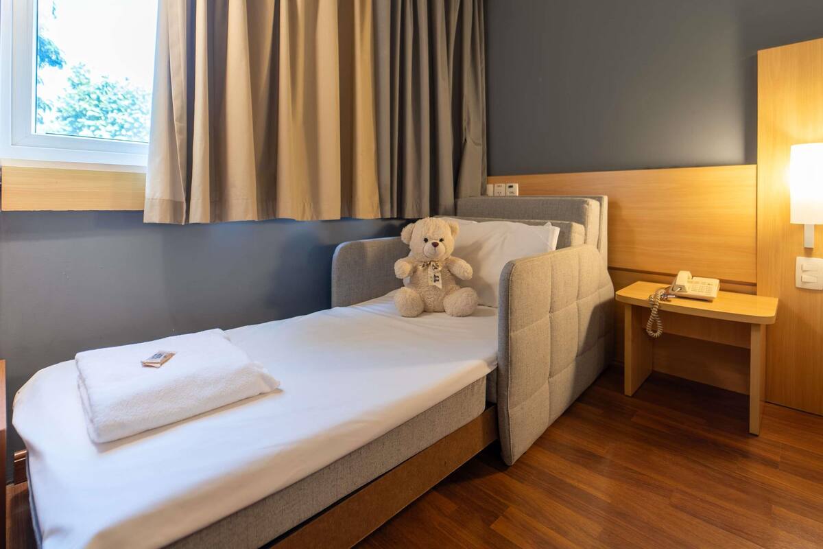 A single bed with a white mattress and a beige teddy bear on it. There is a bedside table with a lamp and a phone. The room has a window with curtains, and the walls are painted in a neutral color.