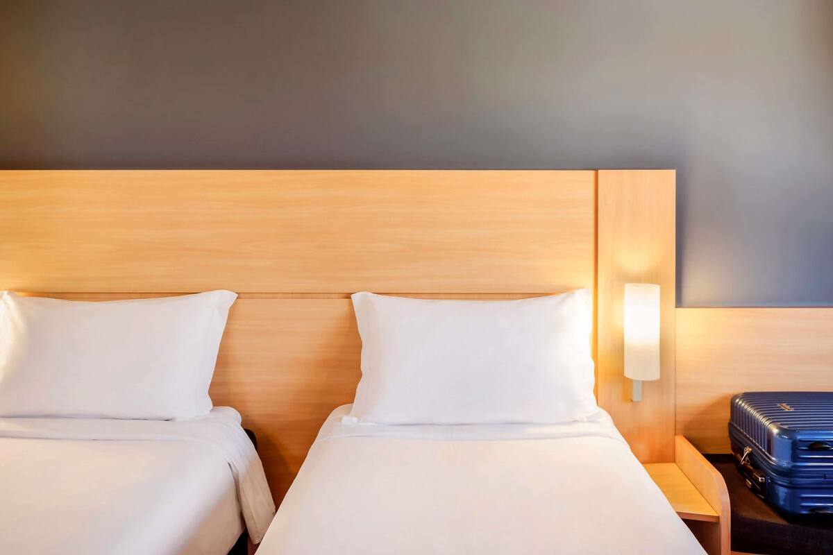 A hotel room with two beds, a nightstand, a lamp, and a suitcase.
