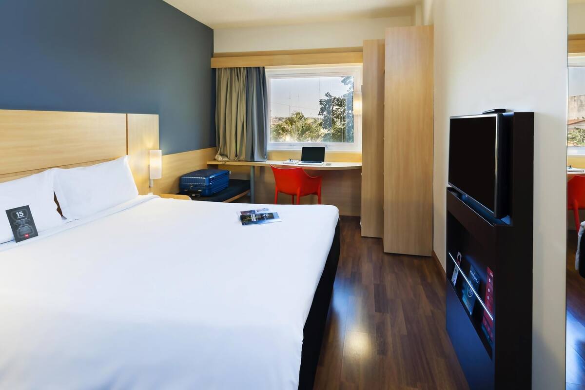 A hotel room with a bed, a desk, a TV, and a window with curtains.